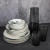 A stack of plates and bowls, a pitcher, and two glasses are arranged on a dark surface against a textured gray background. Anteprima dell’immagine prodotto