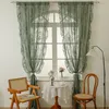 A room interior with a window dressed with green lace curtains, a small round table with a lace tablecloth, a chair, and a floor lamp. Produktbild-Vorschau