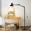 A black adjustable floor lamp is positioned next to a wooden desk with a framed picture and a small green van decoration on it. A grey chair is in the foreground. Náhľad obrázku produktu