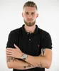 A man wearing a black polo shirt and a silver watch, posing with his arms crossed. Produktbild-Vorschau
