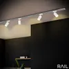 A modern kitchen or living area with track lighting above, featuring dark walls and a wooden shelf. Produktbild-Vorschau