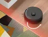 A black robot vacuum cleaner is shown on a wooden floor, cleaning near a rug. Red grid lines indicate its cleaning path and a white arrow shows its movement direction. Produktbild-Vorschau