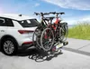 A white car with a bicycle rack attached to the rear, carrying two bicycles, parked on a paved surface near a beach. Produktbild-Vorschau