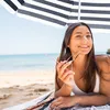 A woman is lying on a beach under a striped umbrella, holding sunglasses. Voir les images du produit