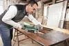 A man is sanding a wooden table with a Bosch belt sander in a workshop. Podgląd zdjęcia produktu