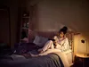 A woman is reading on a tablet in bed at night with her dog lying next to her. Produktbild-Vorschau