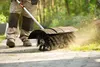 A person is using a powered sweeper on a paved walkway next to grass. Produktbild-Vorschau