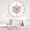 A wall clock with a flower design is mounted on a wall above a bed with pink and blue pillows and blankets. Produktbild-Vorschau