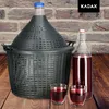 A large glass carboy in a protective basket, a bottle of red liquid, and two glasses of red liquid are arranged on a wooden surface in front of a brick wall. A brand logo is visible in the top right corner. Anteprima dell’immagine prodotto