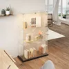 A clear glass display cabinet with LED lighting, showcasing books and decorative items, placed in a well-lit room with wooden flooring and sheer curtains. Produktbild-Vorschau