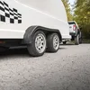 A white trailer with a checkered flag decal is being towed by a pickup truck on a gravel road, with trees in the background. Produktbild-Vorschau