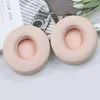 Two pink ear pads for headphones are displayed on a light gray and white surface, with a book and a camera in the background. Náhled obrázku produktu