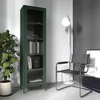 A dark green metal cabinet with glass doors stands next to a large potted plant. A modern chair and a floor lamp are also visible in the room. Produktbild-Vorschau