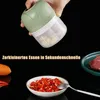 A hand holding a small electric food chopper filled with garlic cloves, with chopped chili peppers and a piece of meat visible in the foreground and background. Produktbild-Vorschau