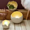 Three decorative egg-shaped bowls are displayed on a wooden surface. One bowl contains greenery and fairy lights, another holds a lit candle, and the smallest also holds a lit candle. A bronze deer figurine stands to the left. Produktbild-Vorschau