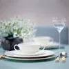 A table setting with a white cup and saucer, plates, cutlery, a glass of water, and a vase of white flowers. Produktbild-Vorschau