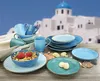 A collection of blue and teal ceramic dishes, cups, and bowls are arranged on a wooden table in front of a blurred background of white buildings with blue domes, suggesting a Mediterranean or Greek island setting. Produktbild-Vorschau