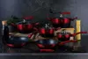 A collection of red and black cookware set with red handles, arranged on a dark countertop with a dark textured background. A bottle of dark liquid, a bunch of spaghetti, and some kitchen utensils are also visible. Voir les images du produit