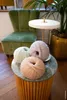 Three balls of yarn are placed on a marble-topped table in front of a green velvet chair and a potted plant. Produktbild-Vorschau