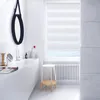 A modern bathroom with a white bathtub, a round mirror, and a window with blinds. Podgląd zdjęcia produktu