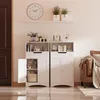 Two white storage cabinets are shown side-by-side in a room with a brown door and a brown leather sofa in the background. Náhled obrázku produktu