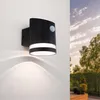 A black outdoor wall light with a motion sensor is mounted on a white wall, with trees and a cloudy sky visible in the background. Produktbild-Vorschau