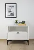 A white nightstand with a drawer and an open shelf, decorated with a framed picture and small decorative items on top. Podgląd zdjęcia produktu