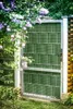 A green privacy screen attached to a metal gate in a garden setting with lilac flowers. Produktbild-Vorschau