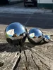 Three polished stainless steel garden spheres of different sizes are placed on a weathered wooden surface. The spheres reflect a building, trees, and a person taking the photo. Náhled obrázku produktu