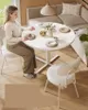 A woman sits at a white round dining table, with plates and a vase of flowers on it. Náhled obrázku produktu