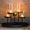 A black metal candelabra with five glass hurricane shades, each holding a lit tea light candle, set against a textured concrete wall. Náhled obrázku produktu