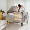 A woman is looking into a baby crib that is set up in a living room. Produktbild-Vorschau