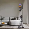 A Dyson vacuum cleaner and its accessories are neatly organized on a stand in a modern living room, next to a sofa and coffee table. Produktbild-Vorschau