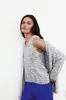 A woman wearing a knitted top and cardigan with blue trousers, leaning against a white wall. Produktbild-Vorschau