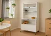 A white kitchen cabinet with glass doors and shelves, placed in a room with a window and a dining table. Anteprima dell’immagine prodotto