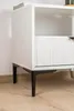 A close-up of the bottom part of a white cabinet with black legs, resting on a wooden floor. Produktbild-Vorschau