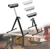 Two adjustable roller stands are shown, one in focus and another slightly blurred with multiple height options indicated by ghosted images. A person's hands are visible in the lower right corner, suggesting the product's use in a workshop setting. Náhled obrázku produktu