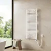 A white electric towel radiator is mounted on a cream-colored tiled wall in a bathroom. A wooden stool with a folded towel and a brush sits in front of the radiator. A large window with a view of greenery is to the left. Podgląd zdjęcia produktu