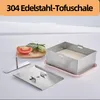 A stainless steel tofu press with its components, a slice of tomato, and some greens on a wooden board, arranged on a white surface. Produktbild-Vorschau