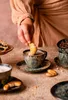 A person is dipping a pastry into a cup of coffee, with other pastries and coffee cups on a textured table. Podgląd zdjęcia produktu