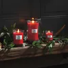 Three red WoodWick candles are arranged on a wooden shelf decorated with greenery and berries. Podgląd zdjęcia produktu