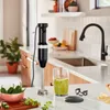 A KitchenAid immersion blender is shown on a kitchen counter next to a glass of blended ingredients, a bowl of jalapeños, and some cilantro. Produktbild-Vorschau