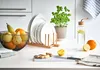 A kitchen counter with various items including a cutting board, ginger, lemons, oranges, a fruit bowl, plates, and a water bottle. Produktbild-Vorschau