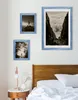 Three framed pictures are displayed on a white wall above a wooden headboard and a bedside table with a lamp. Produktbild-Vorschau