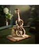 A wooden mechanical cello model is displayed on a stack of books, with a bouquet of flowers in the background. Náhľad obrázku produktu