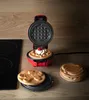 A small red waffle maker with a waffle topped with whipped cream and a raspberry sits on a wooden countertop next to a stove top. Produktbild-Vorschau