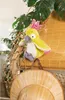 A plush parrot toy is perched on a bamboo blind, with tropical leaves and a straw hat in the foreground. Produktbild-Vorschau