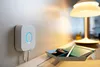 A Philips Hue Bridge is mounted on a wall next to a desk with a lamp and a tray of items. Produktbild-Vorschau