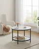 A round coffee table with a marble-look top and black metal legs sits on a rug in a well-lit room. Náhled obrázku produktu