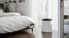 An air purifier is placed in a bedroom next to a bed. Podgląd zdjęcia produktu